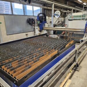 En stor CNC-styrd plasma/gas 1,5 x 3 m Eckert Jantar 2019 Kjellberg SmartFocus 300 med en metallgallerbas inne i en verkstad. Olika verktyg och utrustning är ordnade i bakgrunden.