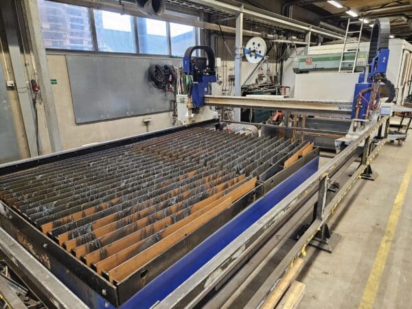 En stor CNC-styrd plasma/gas 1,5 x 3 m Eckert Jantar 2019 Kjellberg SmartFocus 300 med en metallgallerbas inne i en verkstad. Olika verktyg och utrustning är ordnade i bakgrunden.