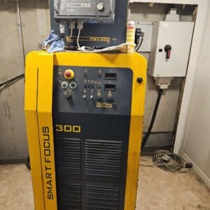 Industrimaskin märkt "CNC-Styrd plasma/gas 1,5 x 3 m Eckert Jantar 2019 Kjellberg SmartFocus 300," med olika knappar, reglage och kablar. Den ligger i verkstadsmiljö med betonggolv och verktyg i närheten.