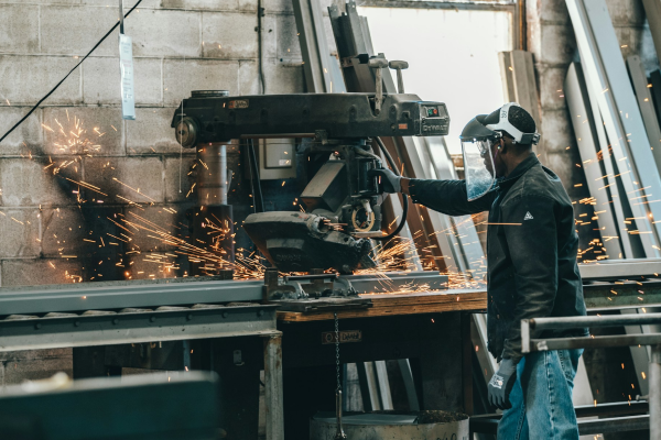 En person som bär skyddsutrustning driver en industrimaskin som producerar gnistor i en verkstad med metall- och betongväggar.