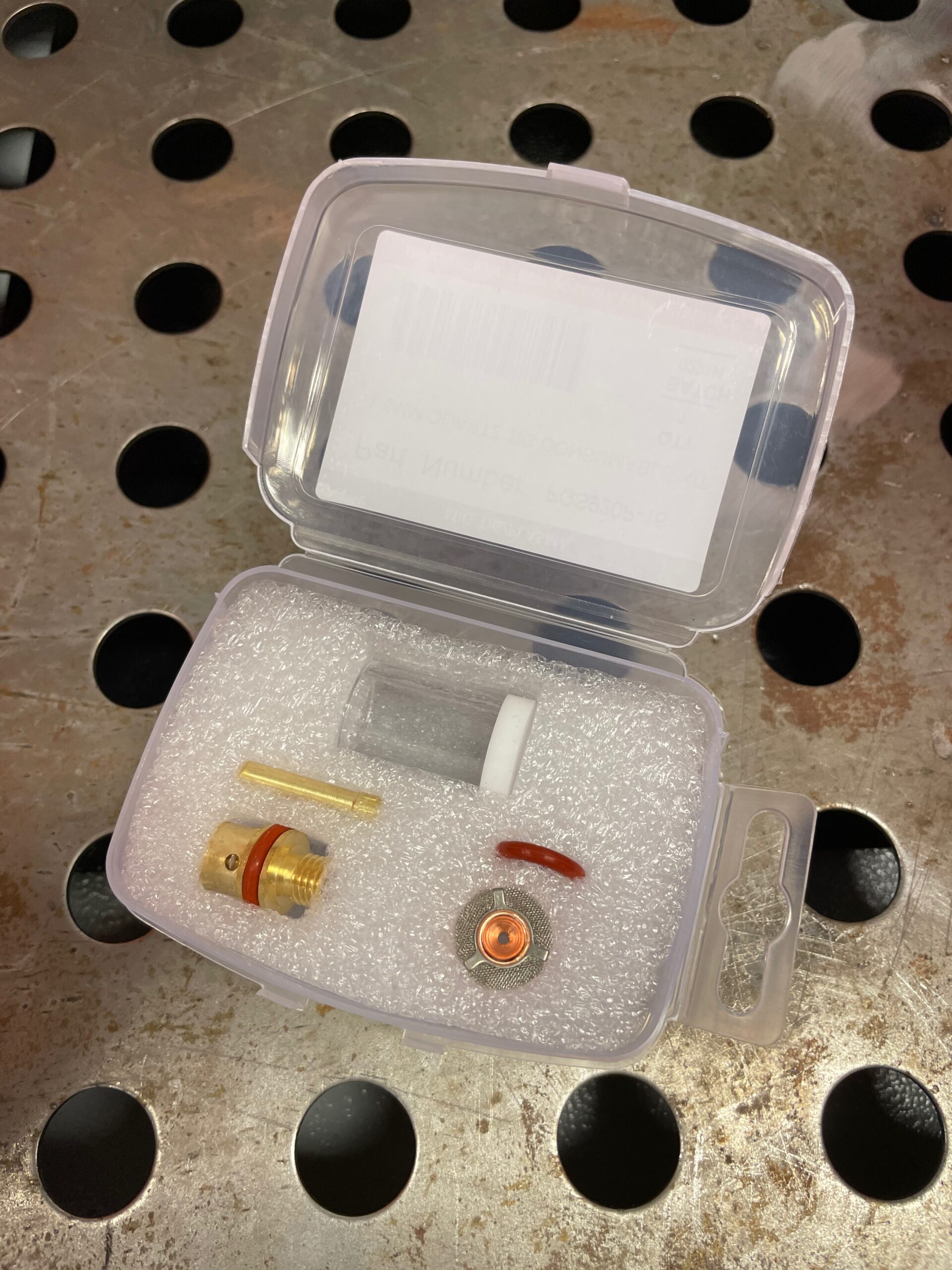 Ett litet genomskinligt plastfodral som innehåller olika metallkomponenter, en gummiring, en kort bit slang och ett litet nätfilter, allt säkert förpackat i skum: Gaskåpa i glas till TIG - Quartz TIG kit - gas saver kit.