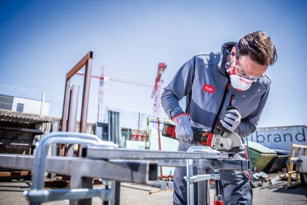 Person som bär skyddsutrustning använder en FLEX Batteridriven vinkelslip med broms 18,0 V, 125 mm LB 125 18,0-EC C för att skära en metallstruktur i en industriell utomhusmiljö. Kranar och byggmaterial syns i bakgrunden.