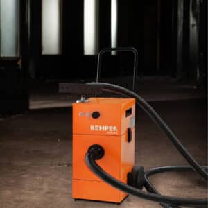 En Högvakuumutsug Kemper Dusty Evo, en orange industrimaskin med hjul och slangar, står på ett betonggolv i ett svagt upplyst rum med stora fönster i bakgrunden.