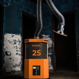 En orange industrimaskin märkt "Kemper mobilt filterutsug SmartFil" står på ett betonggolv, med en flexibel utsugningsslang som sträcker sig uppåt mot en mörk, sliten blå och vit vägg.
